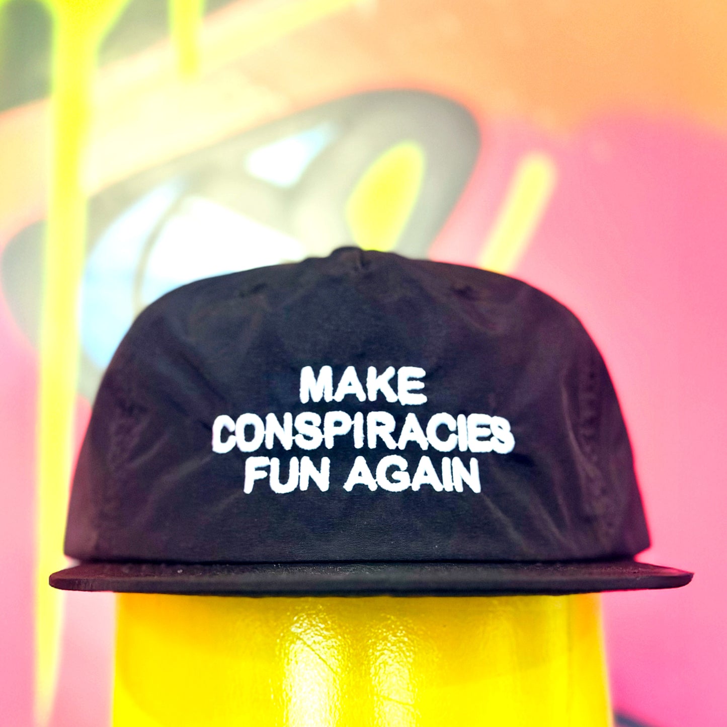 Black cap with 'MAKE CONSPIRACIES FUN AGAIN' text against a colorful, abstract background